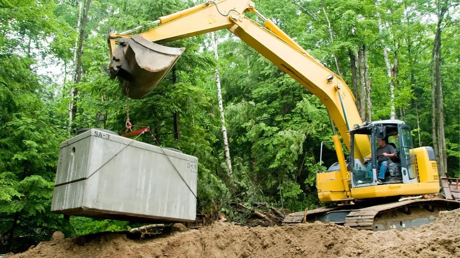 Excavator lowering a concrete septic tank into place on a wooded lot